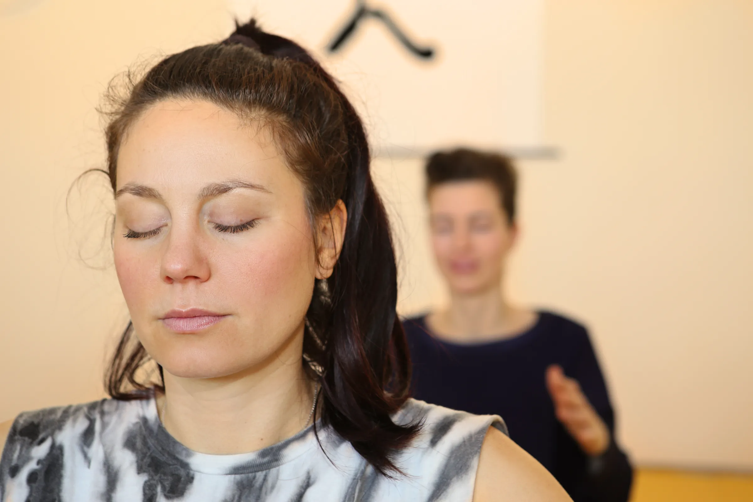 Eine Teilnehmerin meditiert im Yogastudio, eine weitere Person sitzt im Hintergrund.